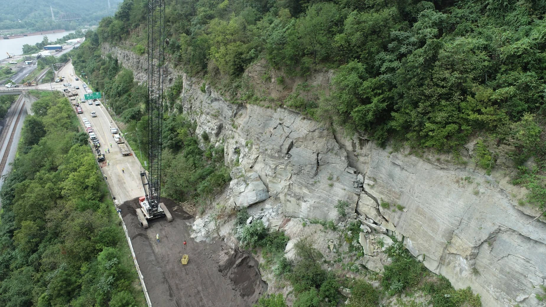 An aerial photo shows a crane and crew working on roadway next to train tracks and a river.